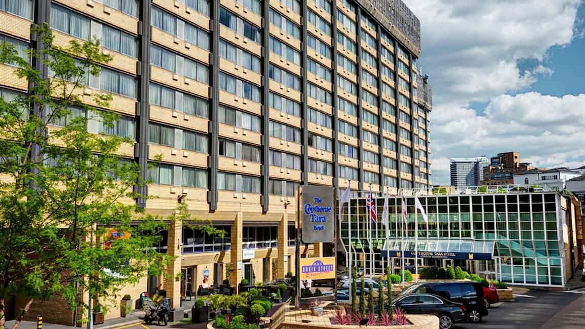 Hotellbygning med flere etasjer, en glassinngang og en parkeringsplass foran. (AI-generert tekst)