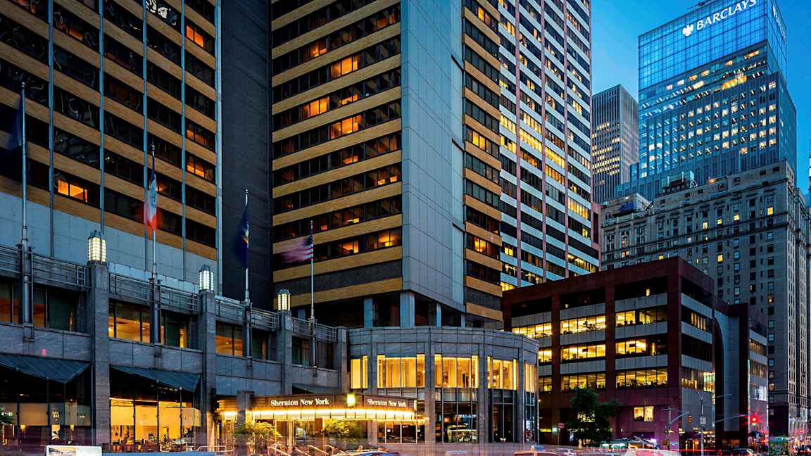 Et bilde av et bymiljø med høye bygninger, hvorav en bygning har teksten "Sheraton New York" ved inngangen. Drosjebiler kan sees på gaten. (AI-generert tekst)