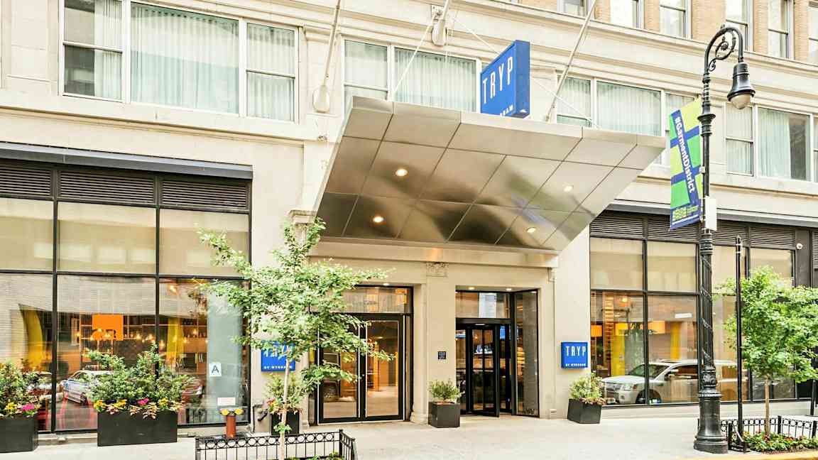 Inngangen til et hotell med skiltet "TRYP". Bygningen har store vinduer og plantekasser. (AI-generert tekst)