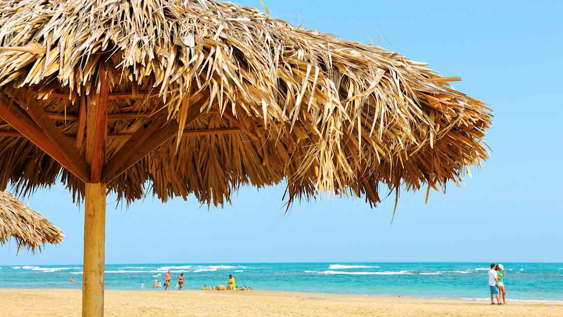 Strand med en parasoll laget av palmeblader i forgrunnen og mennesker på stranden ved havet. (AI-generert tekst)