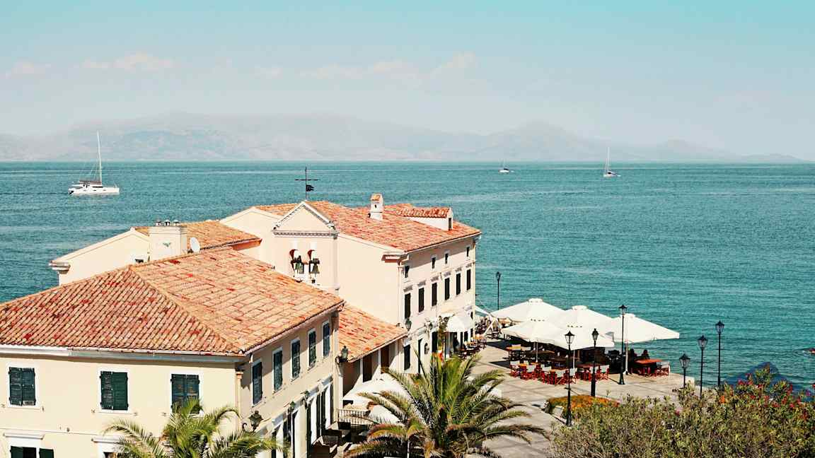 Bilde av bygninger med tegltak ved havet, med palmer og parasoller synlige. (AI-generert tekst)