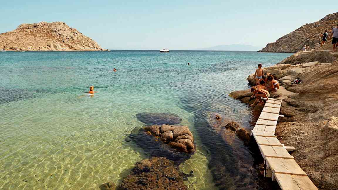 Personer ved en klippe nær en klarblå havbukt med badende. (AI-generert tekst)