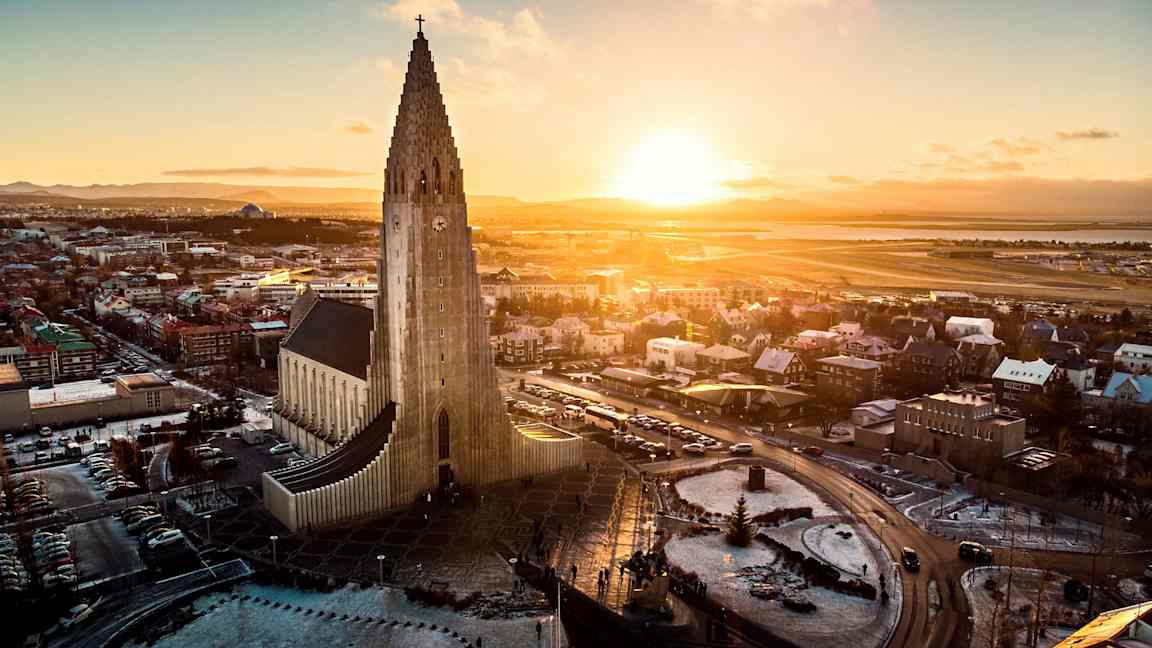 En kirke med moderne arkitektur og en høy tårnstruktur, omgitt av en by og et landskap under en solnedgang. (AI-generert tekst)