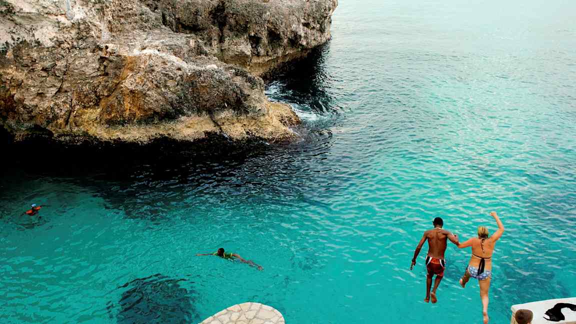 To personer hopper fra en klippe ned i blått vann. (AI-generert tekst)