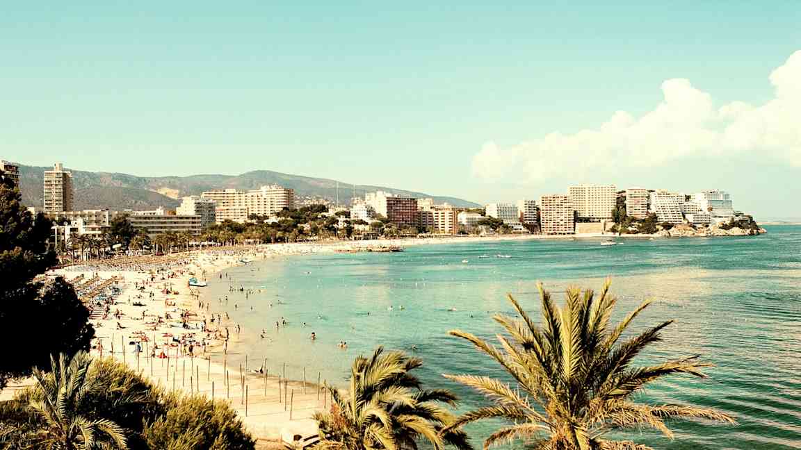 En strand ved havet omgitt av bygninger og palmer. (AI-generert tekst)