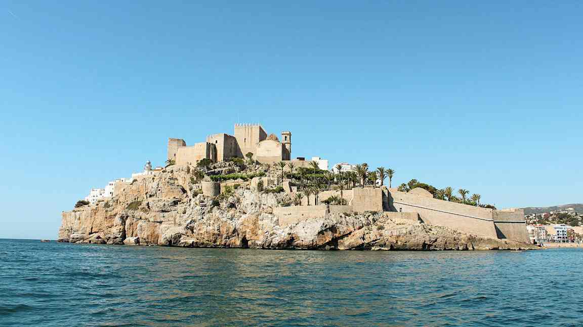 En borg og bygninger på en klippe ved sjøen, omgitt av palmer. (AI-generert tekst)