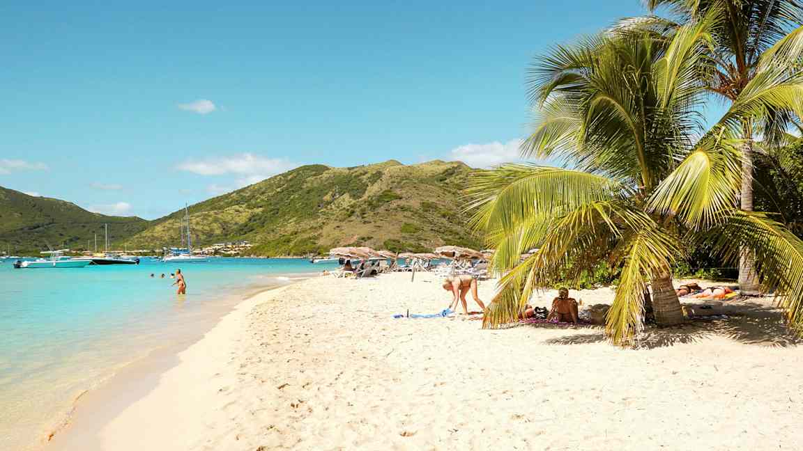 En strand med hvit sand hvor folk slapper av under parasoller. Palme trær ses i hjørnet, og noen bader i det turkise havet. (AI-generert tekst)