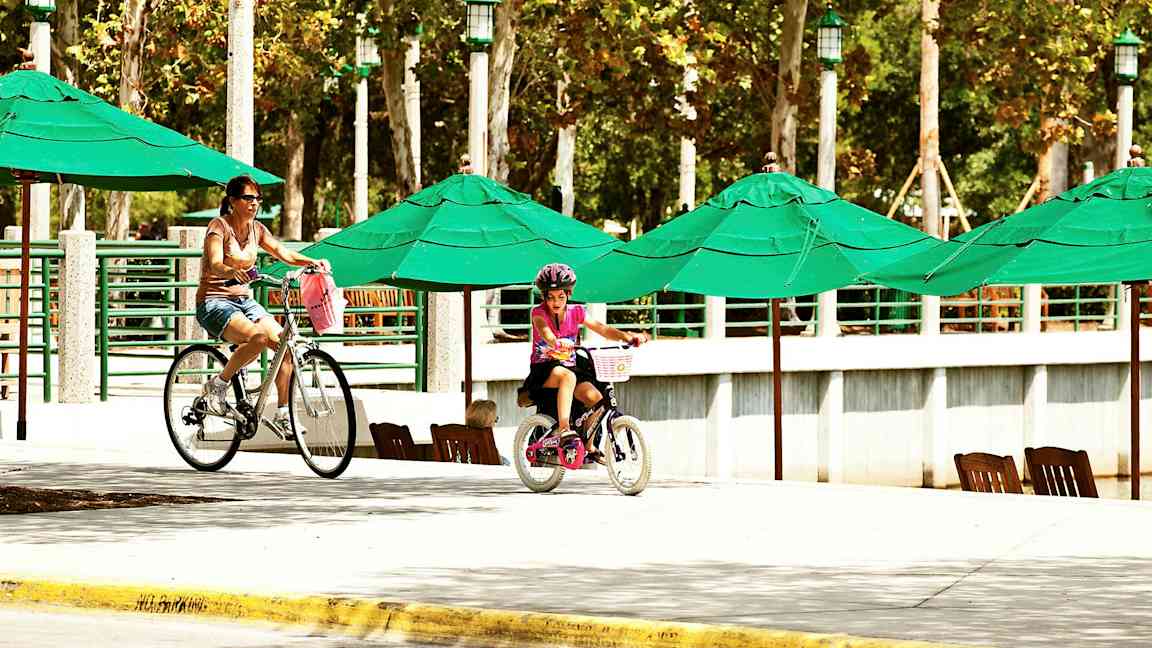 Personer sykler ved et område med grønne parasoller og trerekker i bakgrunnen. (AI-generert tekst)