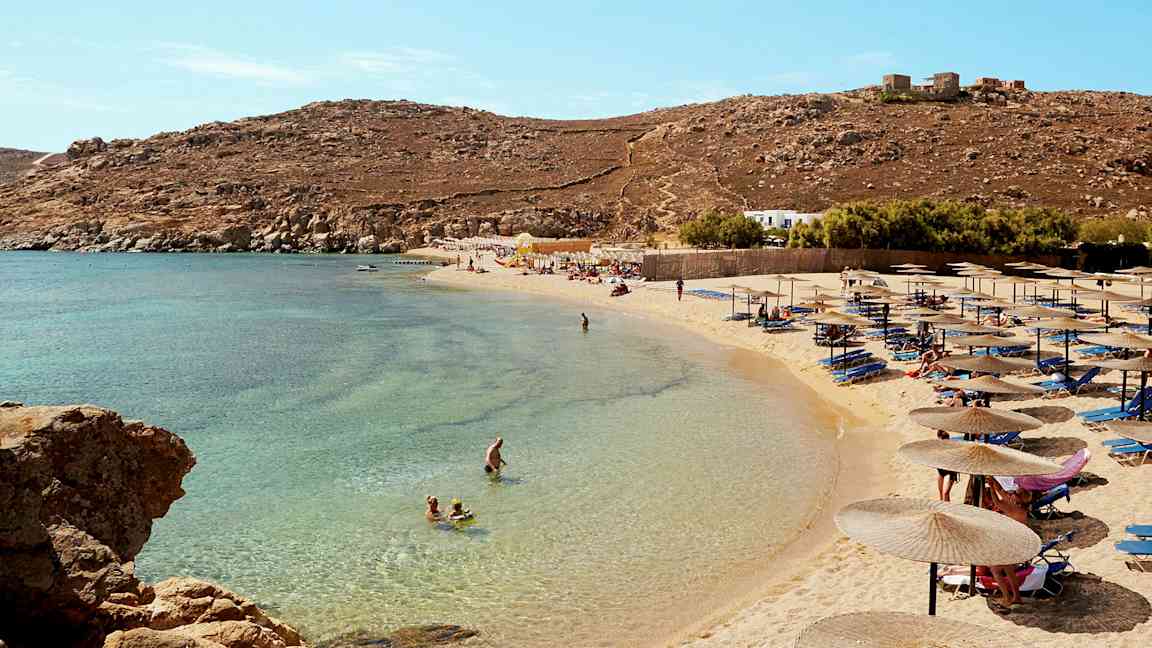 En strand med parasoller og solsenger, folk bader i det klare blå vannet. (AI-generert tekst)