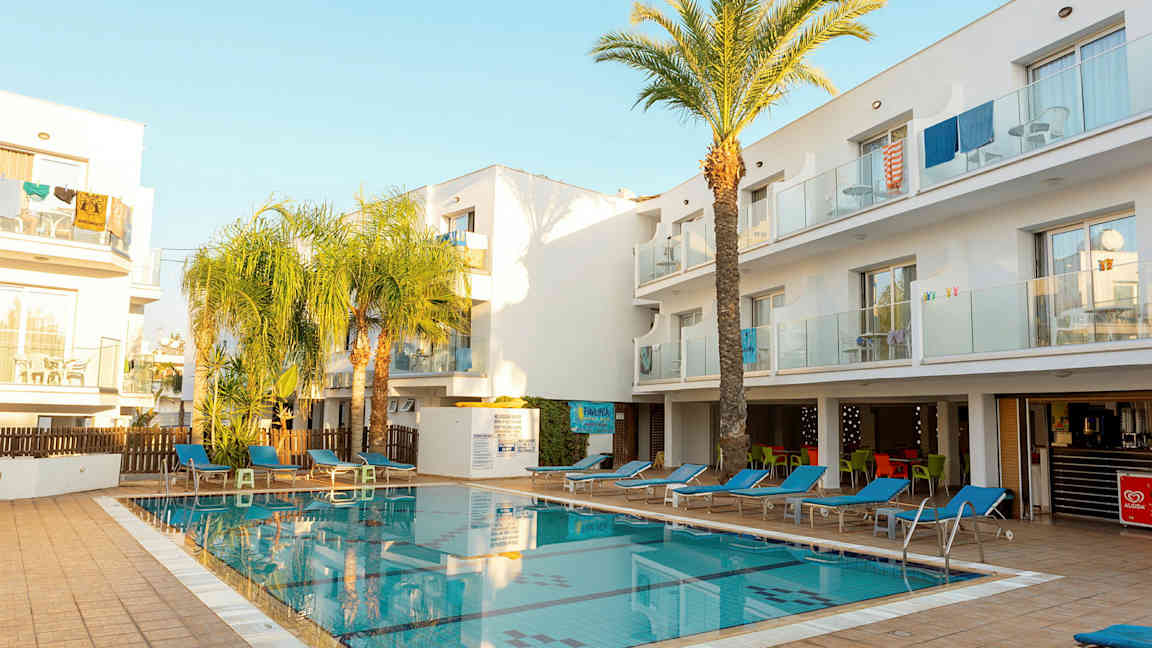 Et utendørsbasseng omgitt av solsenger og hvite bygninger. (AI-generert tekst)