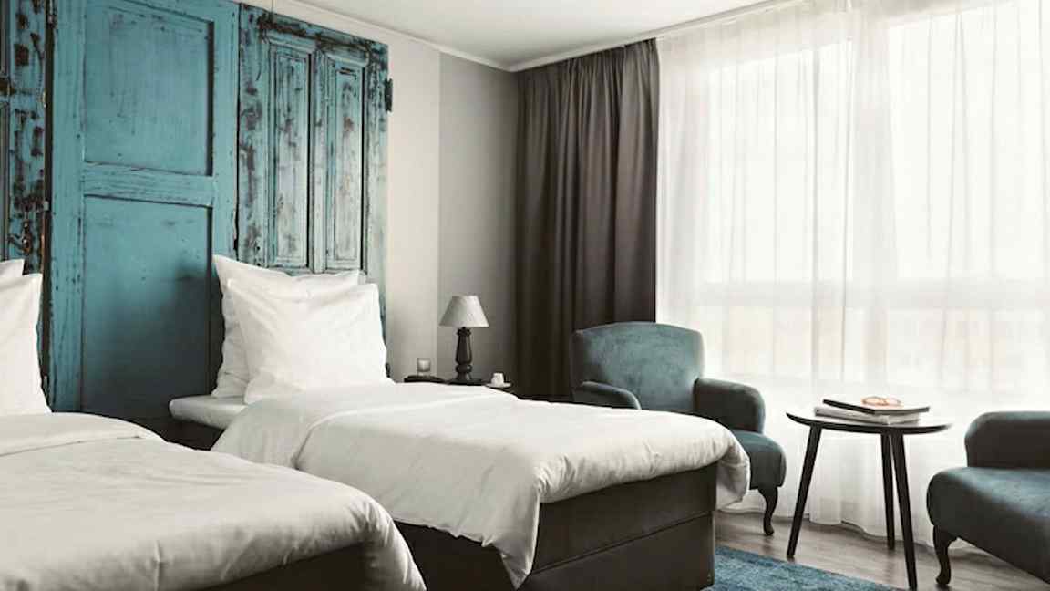 Et hotellrom med to enkeltsenger, blå detaljer på veggen, et bord og lenestoler, samt et stort vindu med hvitt gardin. (AI-generert tekst)