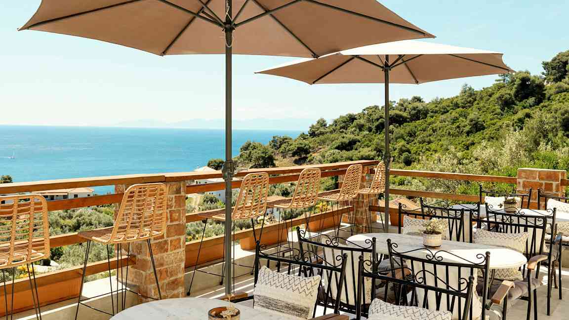 Terrasse med bord, stoler og parasoller med utsikt over havet og frodige omgivelser. (AI-generert tekst)