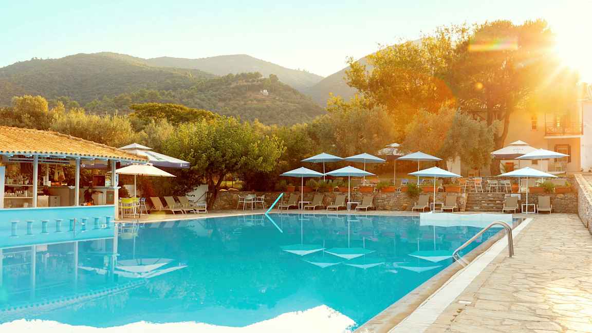 Et utendørs basseng med solsenger og parasoller, omgitt av trær og bygninger i et fjellandskap. (AI-generert tekst)