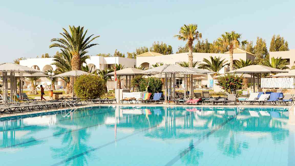 Bilde av et basseng med solsenger og parasoller, omgitt av trær og bygninger. (AI-generert tekst)