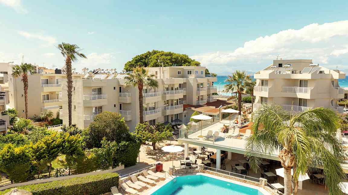 Et hotellområde med basseng, palmetrær og bygninger nær sjøen. (AI-generert tekst)