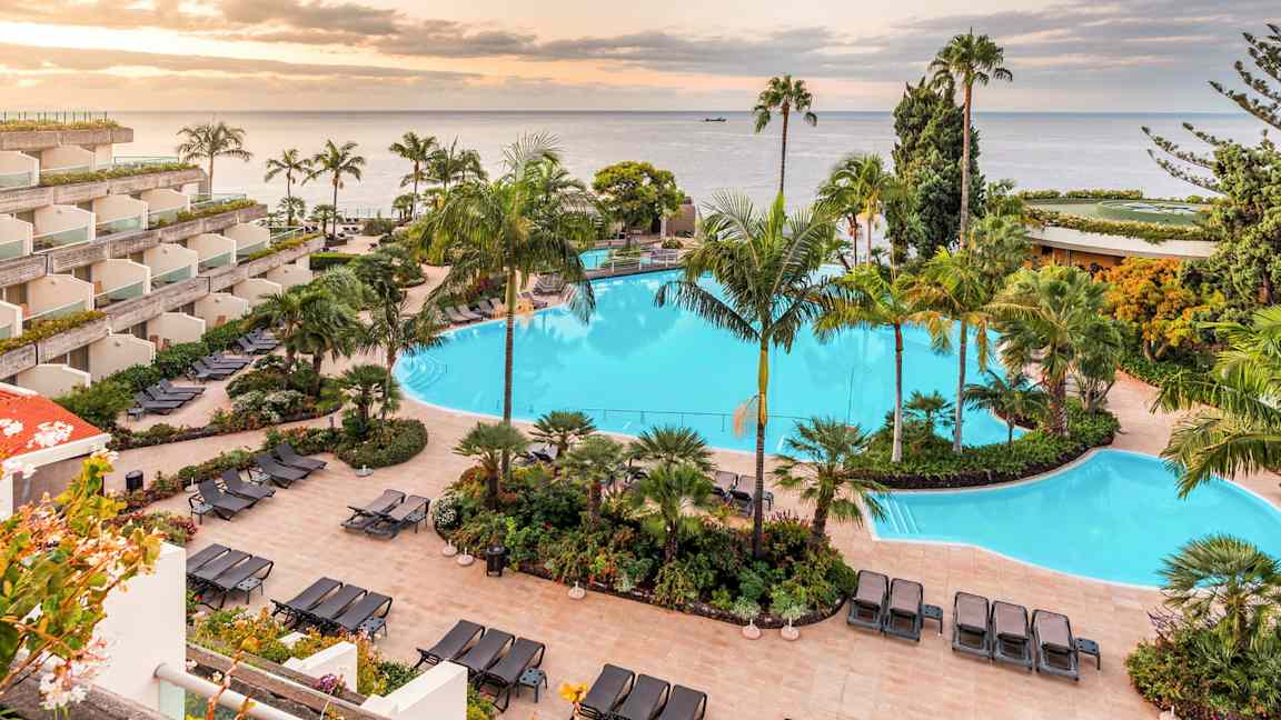 Bilde av et hotellområde med et stort svømmebasseng omgitt av solsenger og tropiske planter. Havet og en solnedgang er synlig i bakgrunnen. (AI-generert tekst)
