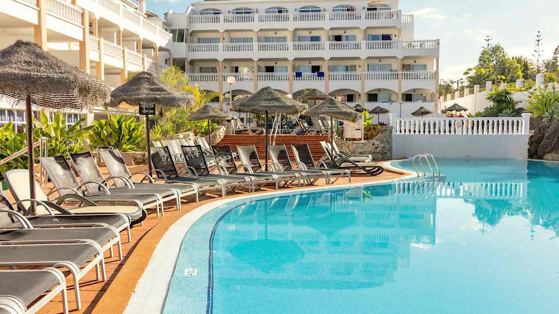 Bilde av en hotellbygning med flere etasjer som ligger ved et basseng. Foran bassenget står solsenger og parasoller. (AI-generert tekst)