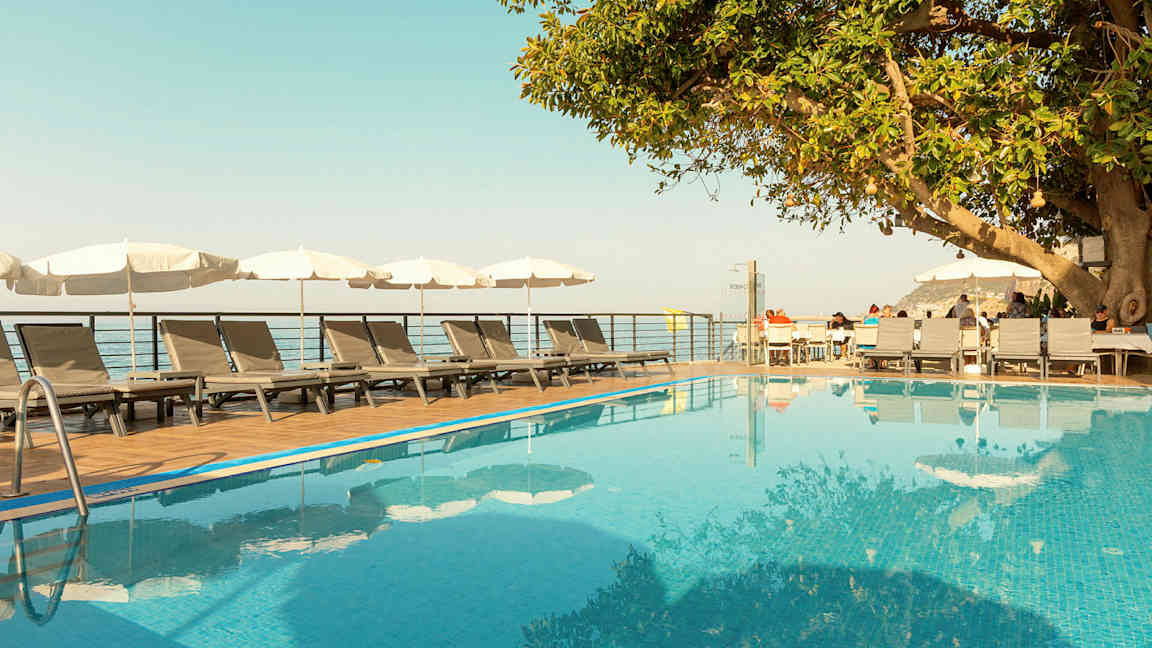 Et utendørs svømmebasseng med solsenger og parasoller ved siden av, samt et tre og en treterrasse med sitteplasser. (AI-generert tekst)