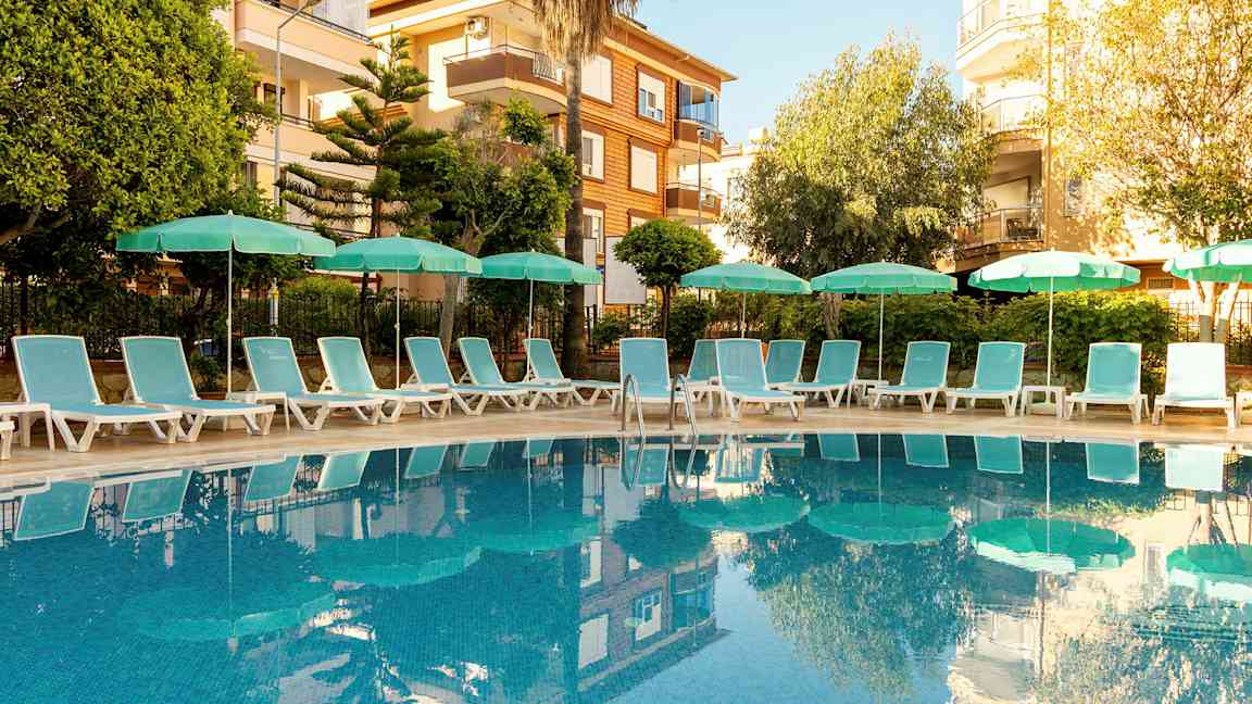 Utendørsbasseng omgitt av solsenger og parasoller, med bygninger i bakgrunnen. (AI-generert tekst)