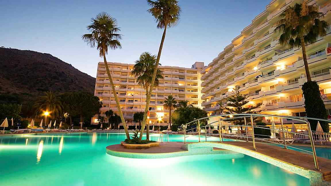 Et bilde av et hotellområde ved skumring, med et opplyst basseng, en bro og høye bygninger i bakgrunnen. (AI-generert tekst)