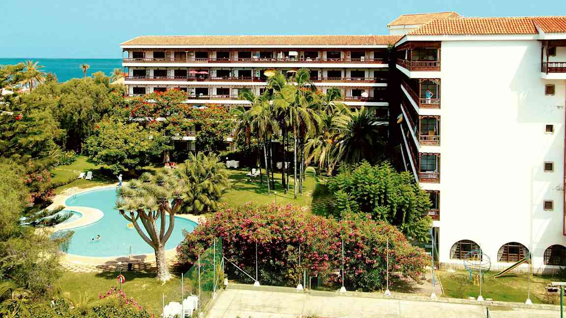 En hotellbygning med balkonger, omgitt av palmer og grøntområder, med et basseng og en tennisbane i forgrunnen. (AI-generert tekst)