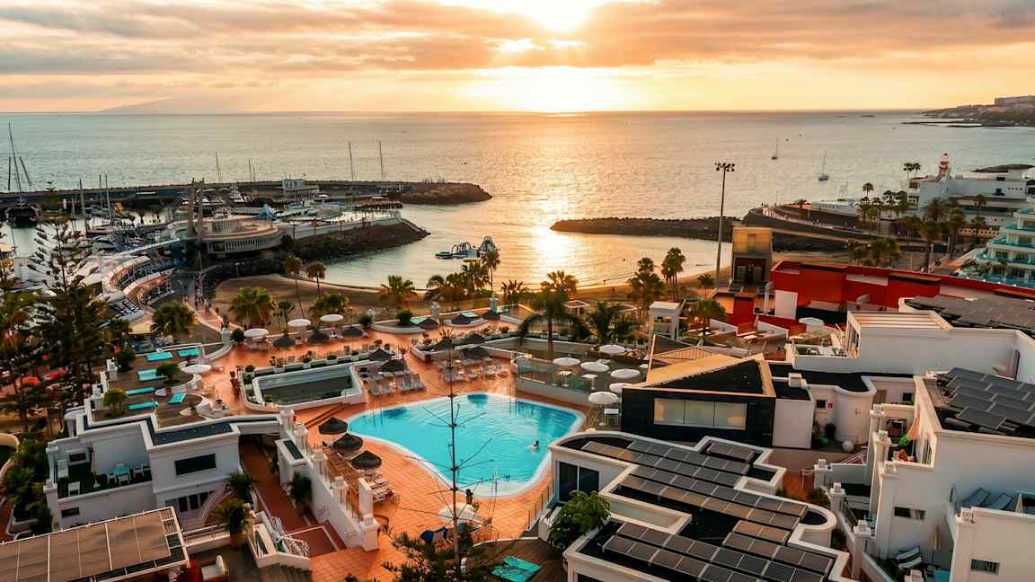 Bilde av en kystoversikt med bassengområde og bygninger i forgrunnen, en havn med båter samt solnedgang over havet. (AI-generert tekst)