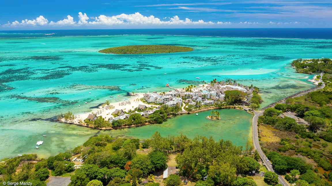 Flyfoto av en strandresort omgitt av turkisblått vann og grøntområder. (AI-generert tekst)