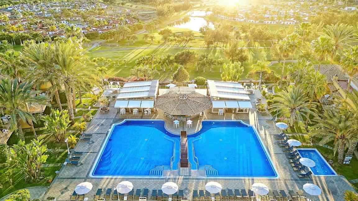 Bilde av to store bassenger ved et feriested, omgitt av palmer og solsenger. (AI-generert tekst)