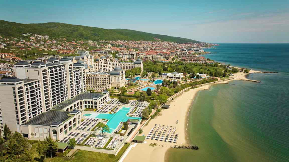 Hotell og bassenger ved stranden med havet og byen i bakgrunnen. (AI-generert tekst)