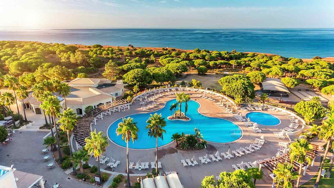 Flyfoto av et hotellområde med store bassenger, solsenger og omkringliggende grøntområde nær stranden. (AI-generert tekst)