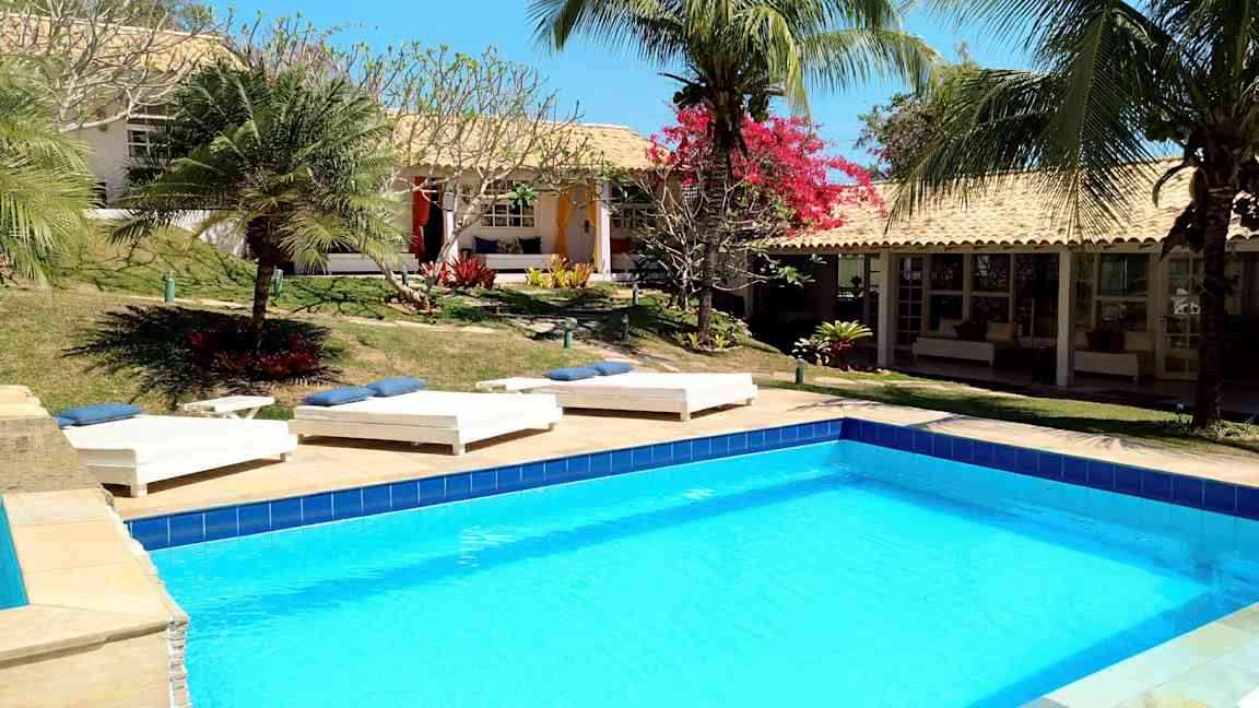 Et basseng omgitt av solsenger og tropiske planter ved en bygning med terrakottatak. (AI-generert tekst)