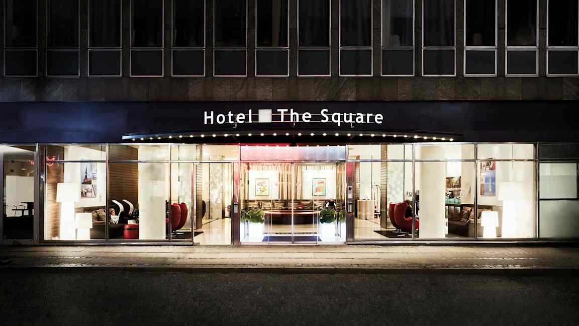 Fasaden og hovedinngangen til Hotel The Square, med glassdører og store vinduer. (AI-generert tekst)