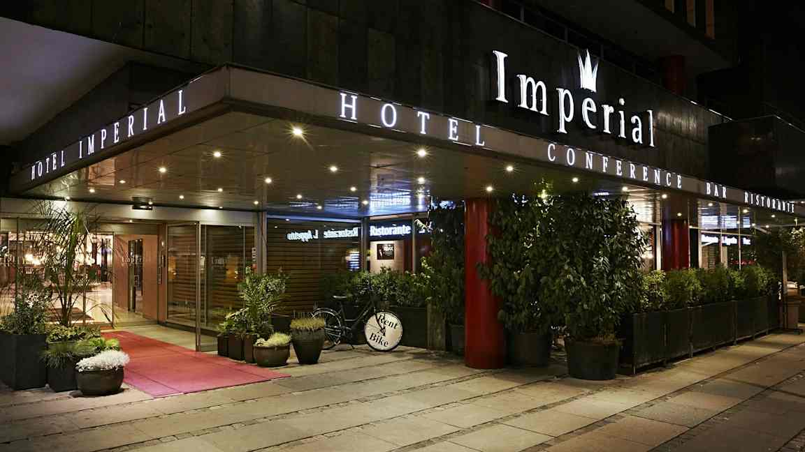 Utvendig bilde av et hotell om kvelden med skilt som viser 'Hotel Imperial', omgitt av planter og en sykkel med skiltet 'Rent Bike'. (AI-generert tekst)