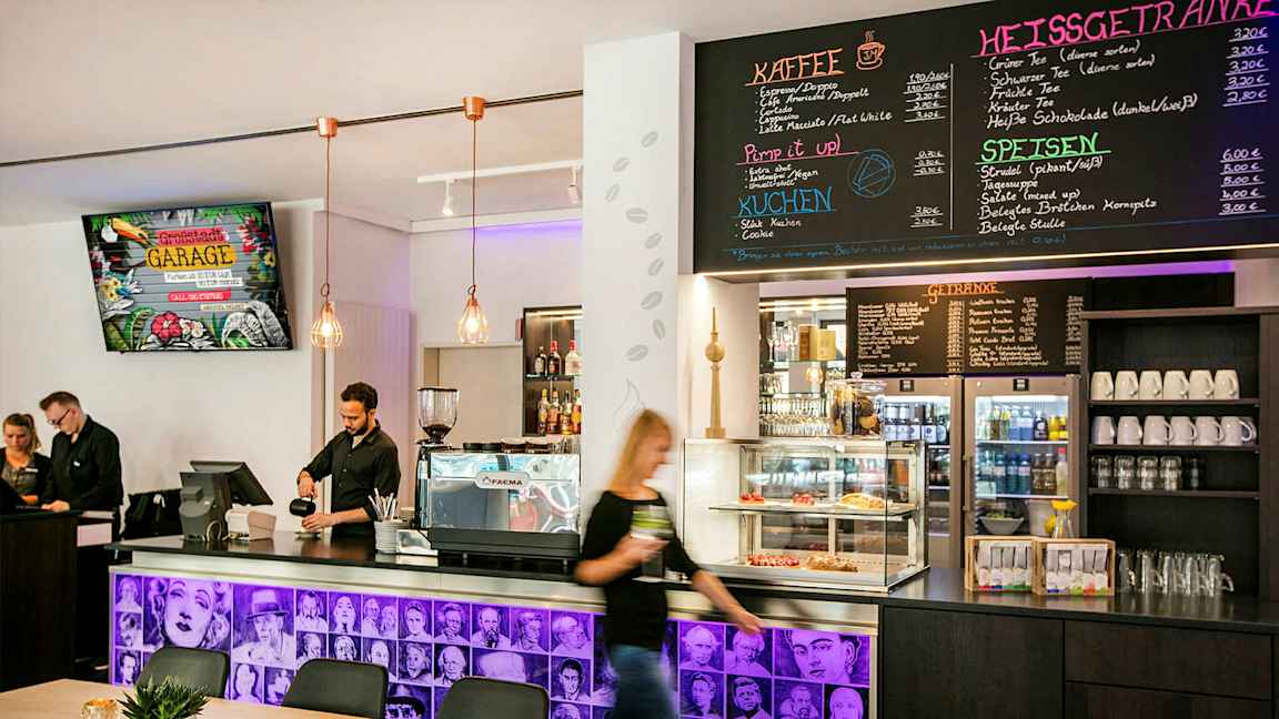 En kaféinteriør med en disk som inneholder kaffemaskin, bakverk i en glassmonter og en meny på veggen over. Ansatte står bak disken. (AI-generert tekst)