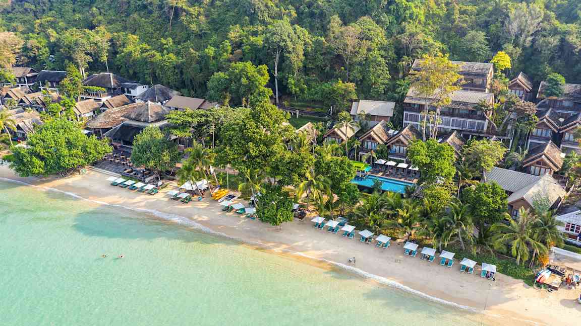 Luftfoto av en strand med solsenger og parasoller samt bygninger omgitt av grøntområder. (AI-generert tekst)