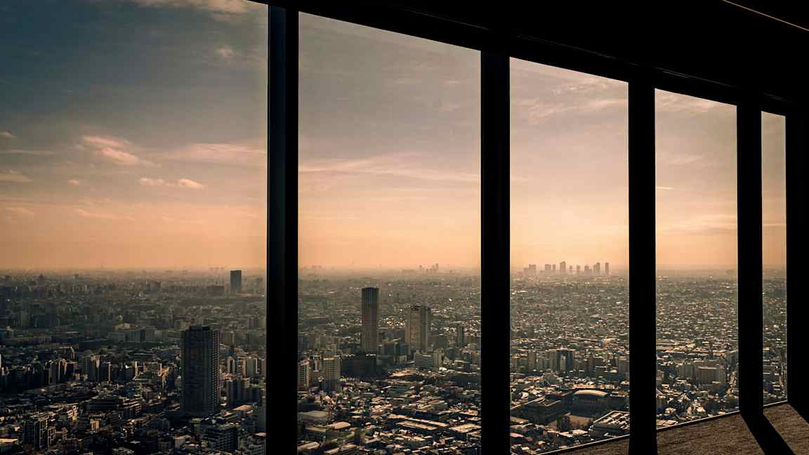 Utsikt over en by gjennom høye vinduer med solnedgang i bakgrunnen. (AI-generert tekst)