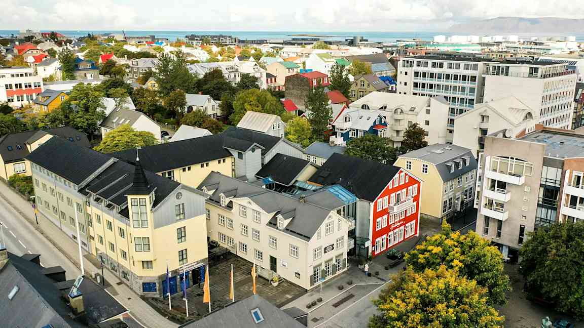 En byutsikt med fargerike bygninger, trær og en vei, samt bakgrunn med hav og fjell. (AI-generert tekst)