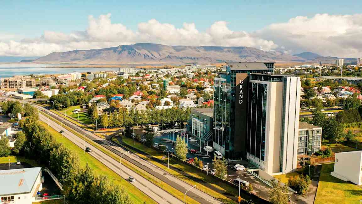 Utsikt over en by med en stor bygning merket Grand i forgrunnen, omgitt av bolighus og grøntområder. I bakgrunnen sees lave fjell og en vannkilde. (AI-generert tekst)