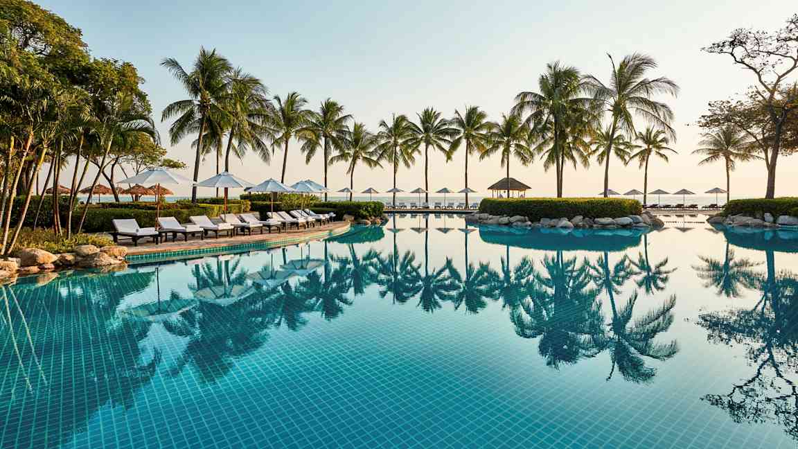 Et utendørs svømmebasseng med klart blått vann, omgitt av solsenger, parasoller og palmer ved havet. (AI-generert tekst)