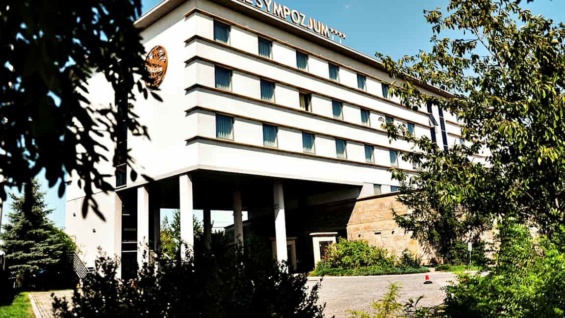 Bygning med flere etasjer, merket med teksten 'Hotel Sympozjum', omgitt av trær og vegetasjon. (AI-generert tekst)