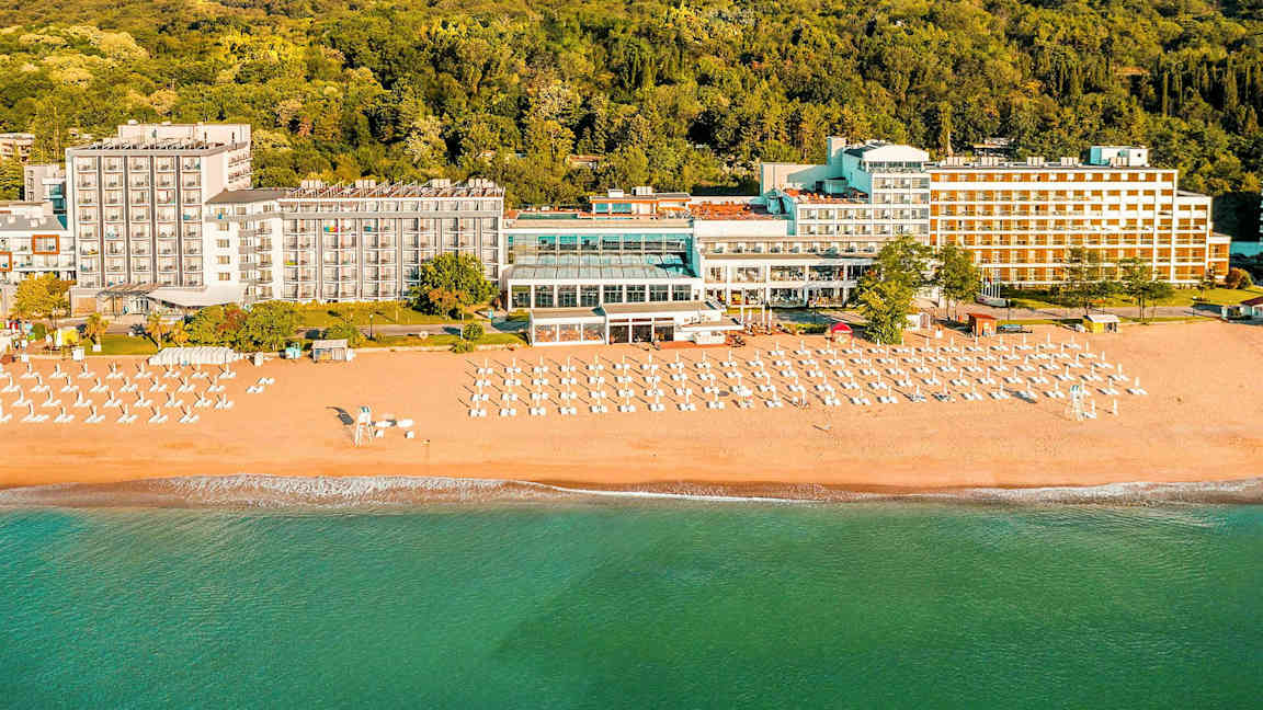 Hotell ved stranden med solsenger og parasoller foran en skog. (AI-generert tekst)