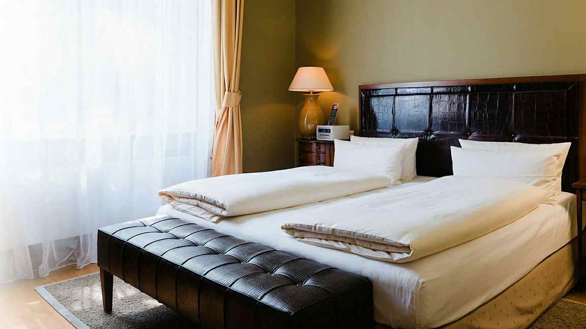 Et elegant hotellrom med en dobbeltseng, puter og lyse sengetøy. Et nattbord med lampe er plassert ved siden av sengen, og et vindu med lette gardiner vises til venstre. (AI-generert tekst)