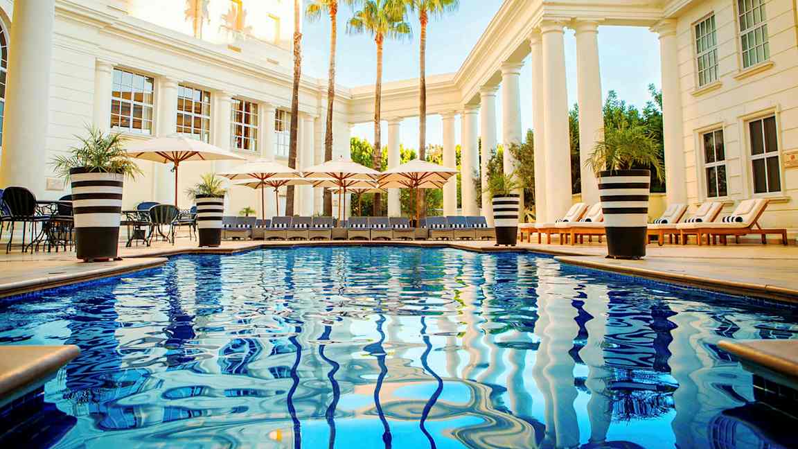 Bassengområde med skinnende blått vann, solsenger og parasoller omgitt av søyler og palmer. (AI-generert tekst)