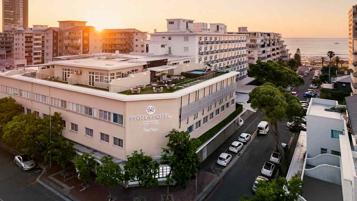 Hotellbygning ved sjøen omgitt av andre bygninger i solnedgang. (AI-generert tekst)