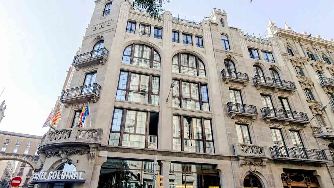 Bygning i klassisk arkitektur med store vinduer og balkonger, pyntet med flagg ved inngangen, inkludert skiltet 'Hotel Colonial'. (AI-generert tekst)