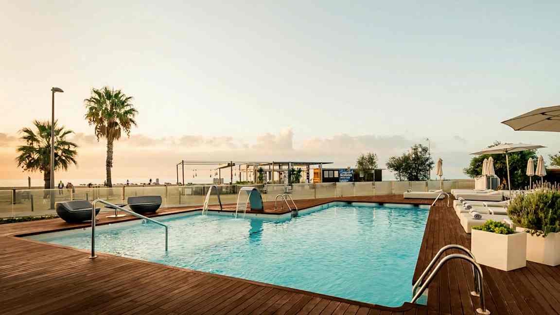 Bildet viser et utendørs svømmebasseng med treterrasse og solsenger ved kanten. I bakgrunnen er noen palmer, en bygning og havet under en lys kveldshimmel. (AI-generert tekst)