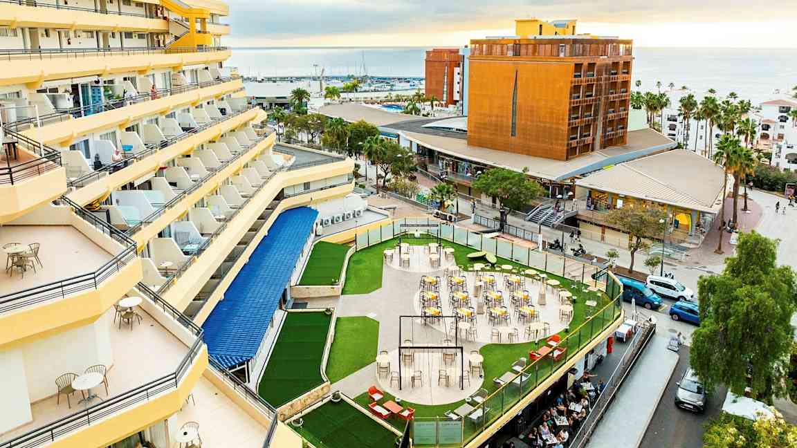 Hotell med gule fasader og balkonger, en terrasse med bord og stoler omgitt av grøntområder, og en utsikt over byen og havet i bakgrunnen. (AI-generert tekst)