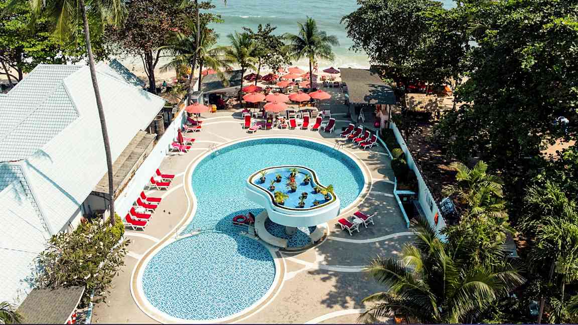 Luftfoto av et basseng på et feriested med solsenger og parasoller, omgitt av tropiske trær og nær havet. (AI-generert tekst)