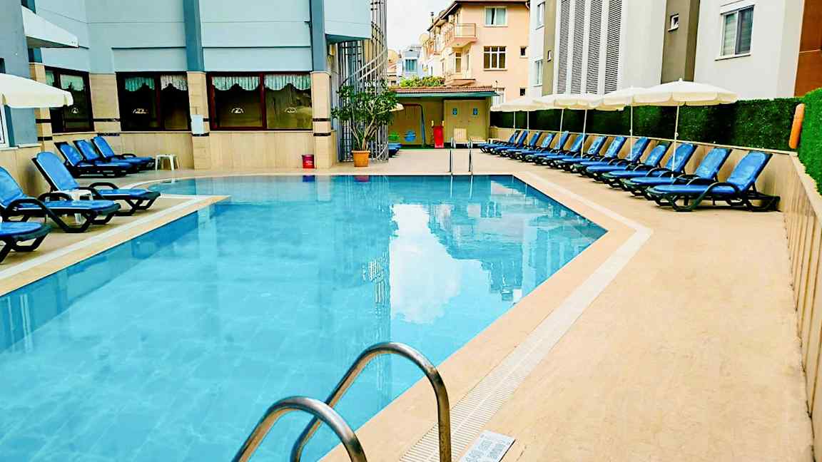 Et utendørs basseng omgitt av solsenger og bygninger. (AI-generert tekst)