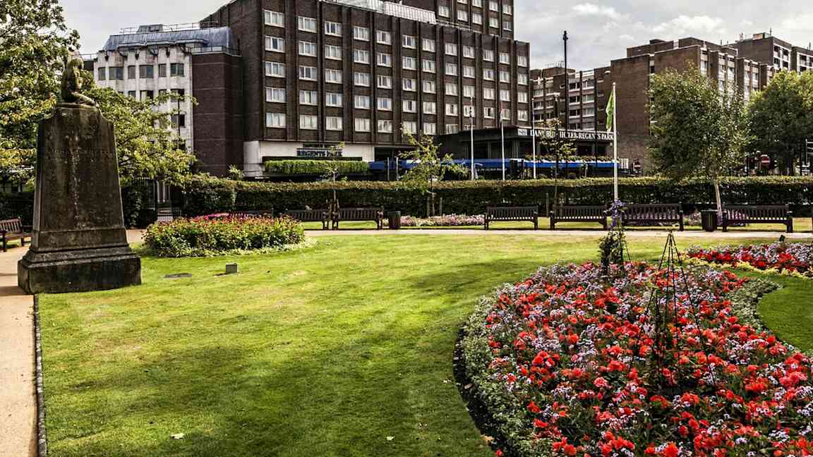 En park med blomster og gressplen foran en bygning. (AI-generert tekst)